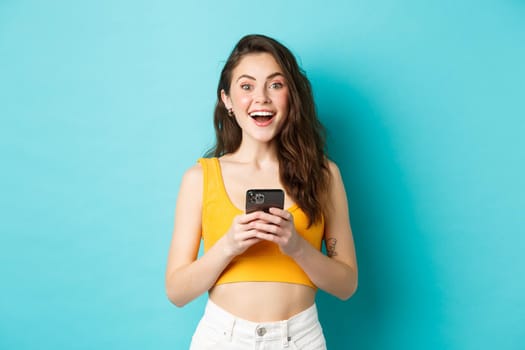 Young excited woman receive great news on phone, holding smartphone, looking amazed at camera with joyful smile, standing against blue background.