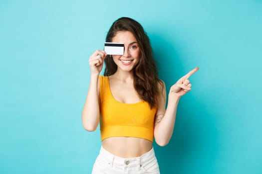 Stylish summer girl with credit card pointing aside, showing your logo on right side copy space, standing against blue background. Copy space