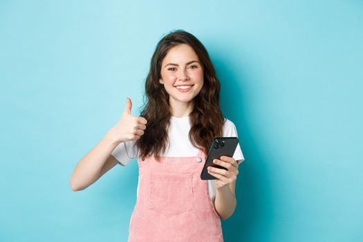 Portrait of beautiful happy girl showing thumb up and holding smartphone, online shopping, making order in internet, looking satisfied, praising good app, blue background.