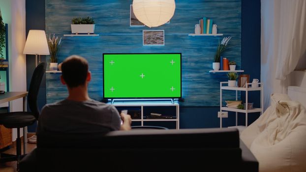 Young man sitting on sofa with green screen on tv in modern living room. Person using television remote with chroma key display for mockup isolated template and blank background design