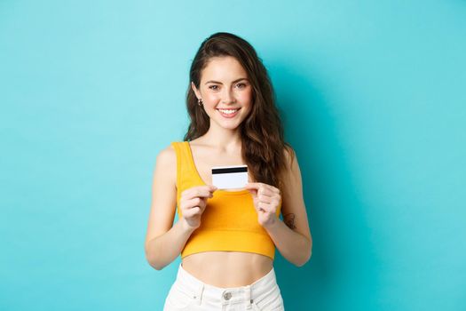 Shopping. Smiling good-looking woman in summer clothes, showing plastic credit card, looking confident, standing over blue background.