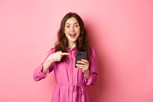 Young woman pointing finger at her phone and smiling, talking about smartphone app, showing something interesting online, standing over pink background.