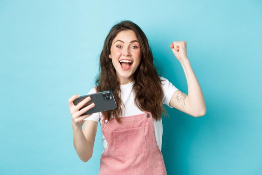 Excited beautful girl winning on mobile phone, holding smartphone and shouting yes with joyful face and fist pump, smiling amazed at camera, blue background.