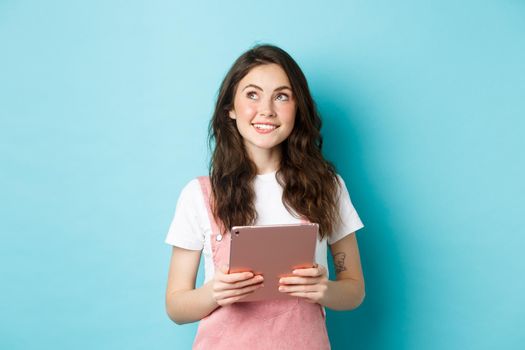 Beautiful dreamy girl smiling, holding tablet in hands and looking at upper left corner logo with thoughtful face, standing over blue background.