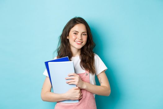 Cute smiling girl student holding notebooks and looking cheerful at camera, studying in college or university, standing over blue background.