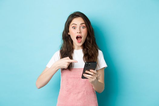 Portrait of surprised attractive female model pointing at smartphone and gasping amazed, checking out online promo offer, showing app, blue background.