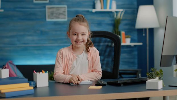 Portrait of young girl writing on notebook with pen smiling while sitting at desk. Little child preparing for school lessons, doing homework and learning. Pupil looking at camera