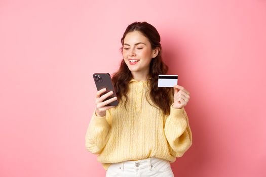 Technology and online shopping. Young pretty lady paying online with credit card, looking at smartphone and smiling, standing over pink background.