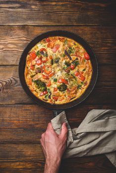 Vegetable frittata with broccoli, red bell pepper and herbs. Hand holds cast iron skillet