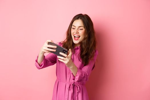 Technology concept. Carefree young woman tilt body, holding smartphone horizontally, playing video game on phone, standing against pink background. Copy space