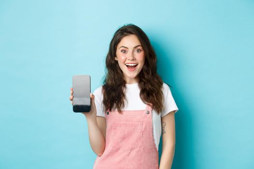 Image of beautiful young woman look surprised and excited, showing empty smartphone screen, your logo or app on mobile display, standing over blue background.