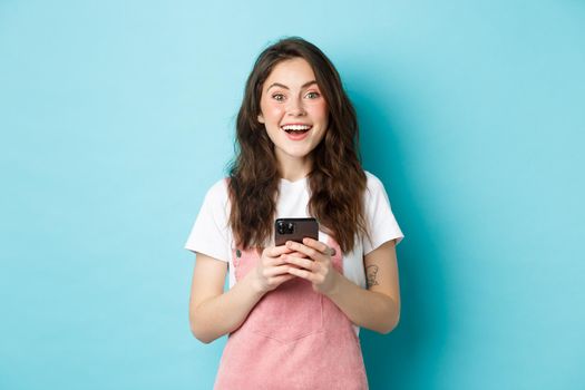 Portrait of excited brunette girl looking amazed at camera while using smartphone, holding mobile phone and smiling cheerful, standing against blue background.