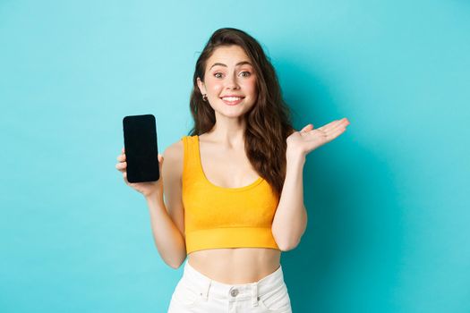 Cheerful young girl introduce your app or store, showing emtpy smartphone screen and smiling at camera, standing against blue background.