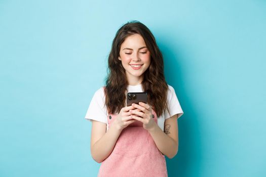 Portrait of young woman using smartphone, reading message or chatting in app, standing over blue background and smiling happy.