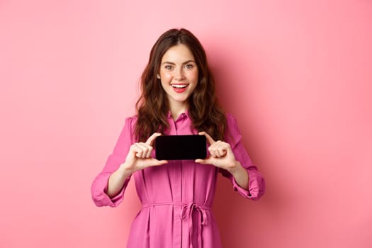 Technology concept. Young attractive woman shows smartphone screen horizontally, introduce online app or promo text, stands against pink background.