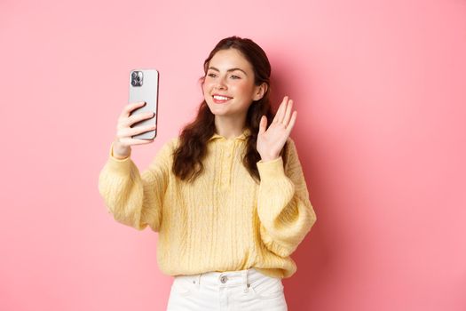 Portrait of friendly girl talking on video chat app, waving hand at smartphone camera, having mobile conversation, standing against pink background.