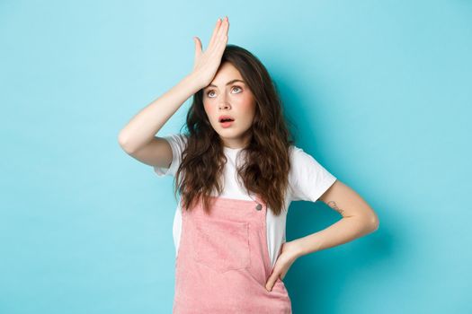 Annoyed and tired young woman in summer outfit, slap forehead and roll eyes, standing over blue background.