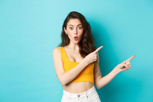 Summer lifestyle concept. Surprised stylish woman showing direction, pointing fingers right, that way gesture, standing over blue background.