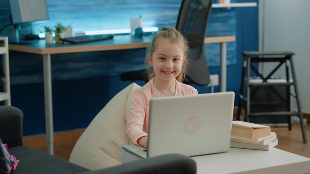 Portrait of young girl working on laptop for school lessons. Schoolgirl using gadget for online remote courses and virtual education while looking at camera and feeling cheerful.