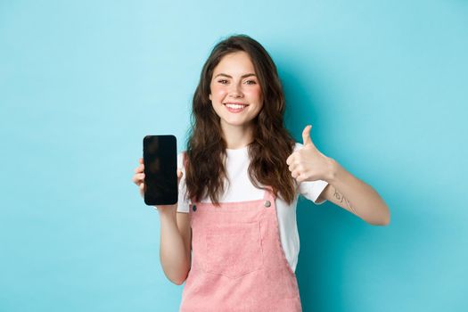 This is good. Smiling cheerful woman showng empty smartphone screen and thumb up to recommend app or online store, standing against blue background.