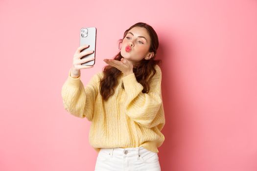 Beautiful flirty girl taking selfie on smartphone, sending air kiss during video chat, standing against pink background. Copy space