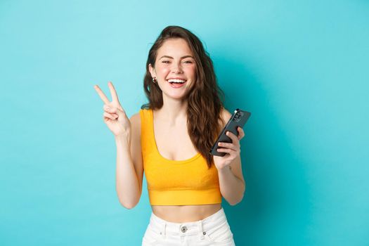 Technology and lifestyle concept. Happy attractive woman laughing, showing v-sign and using smartphone, chatting on mobile phone, standing over blue background.