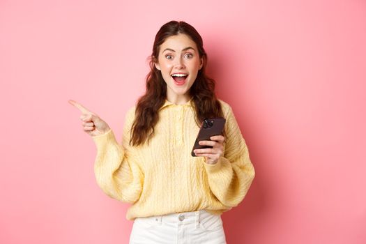 Excited young woman holding mobile phone, pointing finger aside at left copyspace, showing advertisement, found interesting promo online in smartphone, standing against pink background.