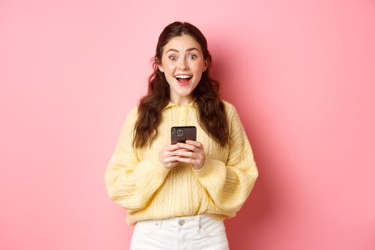 Portrait of girl holding smartphone, looking surprised and gasping excited, found something interesting online, standing against pink background.