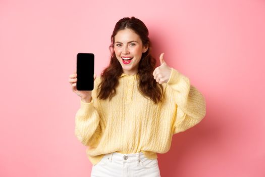 Very good app. Smiling satisfied woman showing thumbs up and empty smartphone screen, recommending app or website, standing against pink background.
