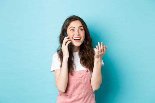 Portrait of cheerful modern girl talking on phone, gesturing and looking happy, smiling at camera, chat with friend on smartphone, standing over blue background.