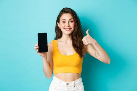 Portrait of stylish modern woman smiling pleased, recommending app, showing you logo banner on empty smartphone screen, making thumb up in approval, praise application, blue background.