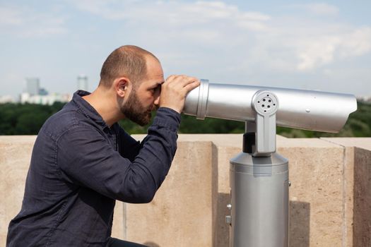 Man tourist standing on building rooftop looking through binocular telescope at metropolitan city admiring beautiful place. Panoramic landscape view from tower observation point.