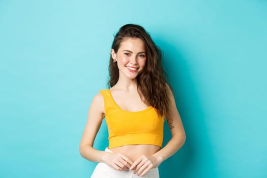 Young stylish woman ready for summer holidays, wearing yellow top and smiling cheerful at camera, standing against blue background.