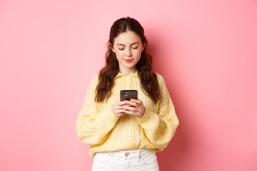 Technology. Young smiling woman chatting on social media, reading message on smartphone, standing against pink background.