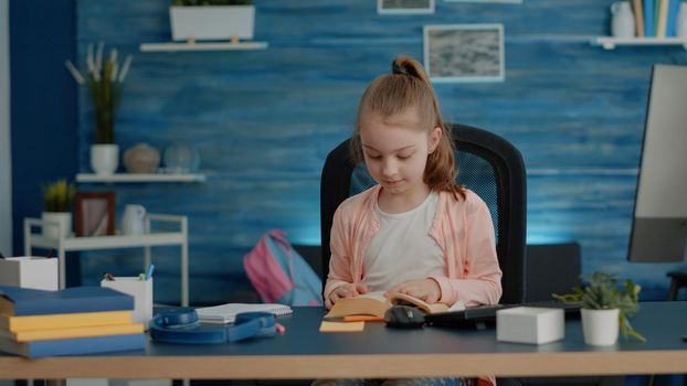 Little child opening book and reading for school classes at desk. Schoolgirl looking at lecture story for online remote literature lesson at home. Young pupil learning for education.