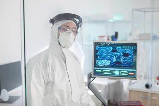 Tired doctor in protection suit against covid-19 looking exhausted at camera behind the glass wall working in danger area. Scientist examining virus evolution using high tech for scientific research