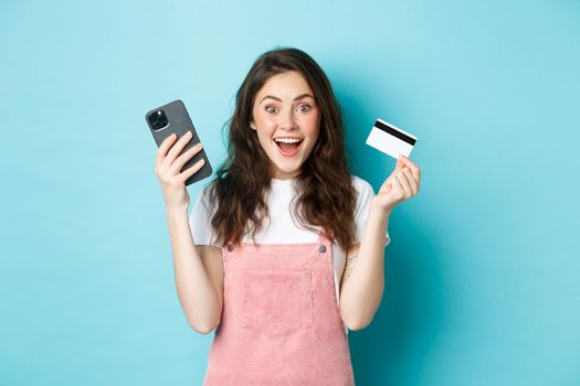 Cheerful girl looks excited at camera, hold smartphone and plastic credit card, standing over blue background.