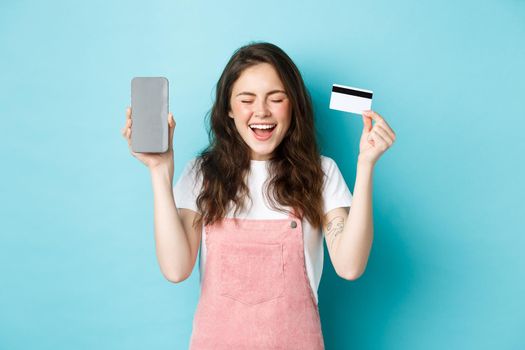 Excited attractive woman scream of joy, showing empty smartphone screen and plastic credit card, raising hands up and smiling happy, standing over blue background.