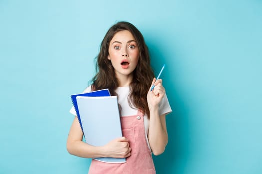 Got an idea. Excited young woman student, holding notebooks and pen, raising hand while having a plan, think-up good solution, standing over blue background.