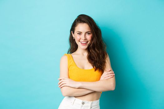 Summer and lifestyle concept. Confident and sassy attractive woman in cropped top looking determined at camera, cross arms on chest and smiling, standing against blue background.