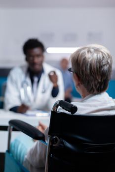 Old patient with disability receiving consultation from black medic in medical cabinet. Invalid senior woman sitting in wheelchair talking to african american doctor about healthcare issues