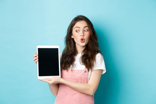 Intrigued glamour girl looking at digital tablet screen while showing it to you, standing amused with online promo offer, demonstrate something on display, blue background.