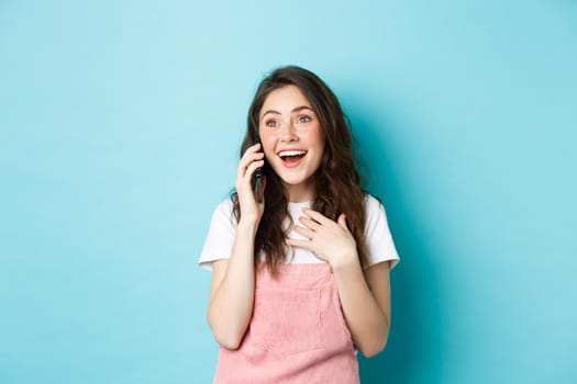 Beautiful girl talking on smartphone and receive surprising news, looking excited and amazed, talking on phone, standing happy against blue background.