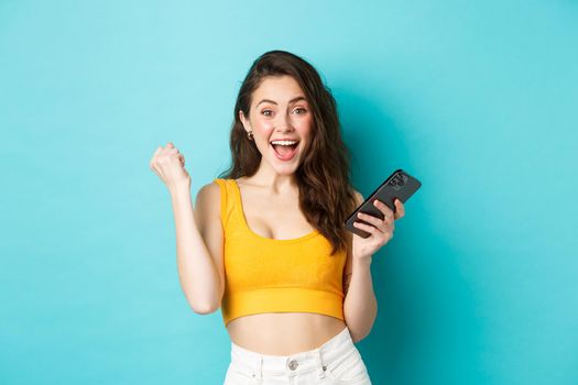 Technology and lifestyle concept. Excited woman winning online, holding smartphone and making yes fist pump gesture, celebrating, achieve goal in app, blue background.