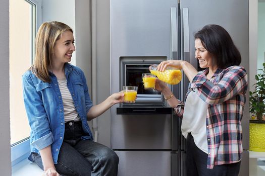 Two middle-aged women drinking orange juice. Smiling female friends talking in the kitchen near the refrigerator. People, food, drinks, diet, rest, communication, lifestyle concept