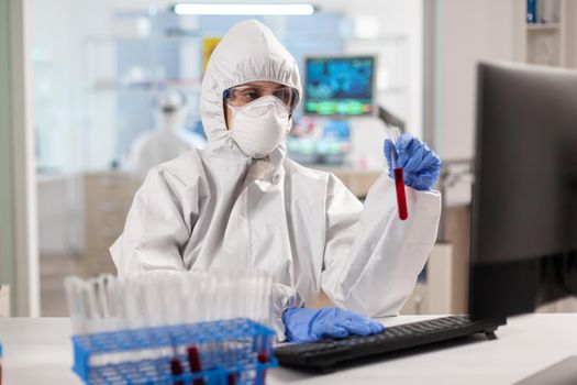 Researcher in coverall holding test tubes with blood sample typing on computer. Scientist examining vaccine evolution using high tech technology for treatment development against covid19 virus.
