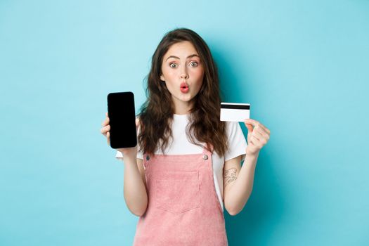 Portrait of young beautiful woman showing plastic credit card and empty smartphone screen, look amused and interested, showing interesting app, standing over blue background.