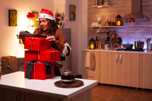 Traditional person carrying gift boxes to counter in festive kitchen at home. Caucasian young adult with santa hat preparing presents for friends and family at christmas eve celebration