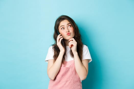 Cute thoughtful girl talking on smartphone and thinking, making choice, order delivery, standing against blue background.