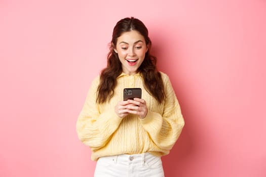 Portrait of excited brunette woman gasping amazed, reading message on smartphone, look surprised at mobile phone, standing over pink background.